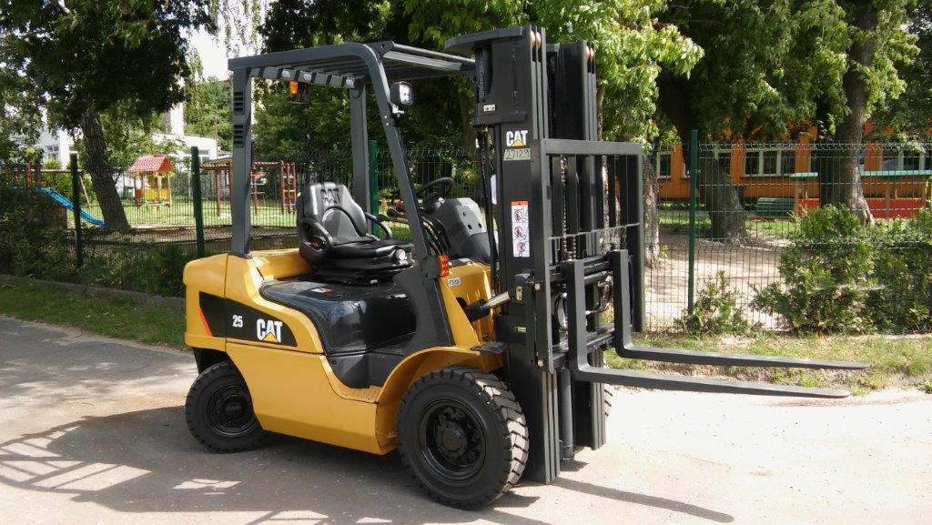 Żółty wózek widłowy marki CAT o udźwigu 2.5 tony, zaparkowany na zewnątrz, z placem zabaw dla dzieci widocznym w tle za ogrodzeniem.