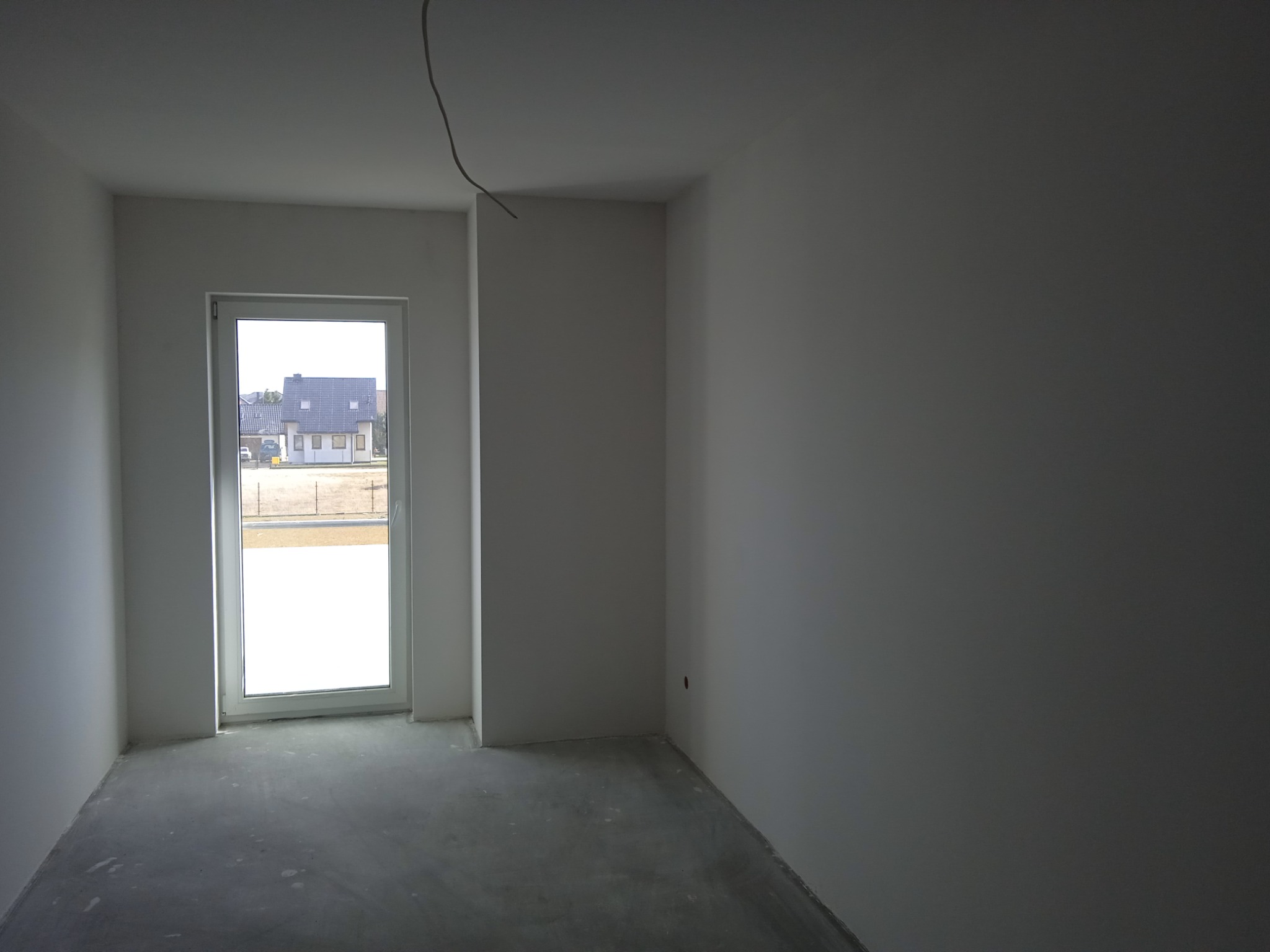 Surowe wnętrze pokoju z oknem, ściany pomalowane na szaro, betonowa podłoga, widoczny kabel wystający z sufitu.