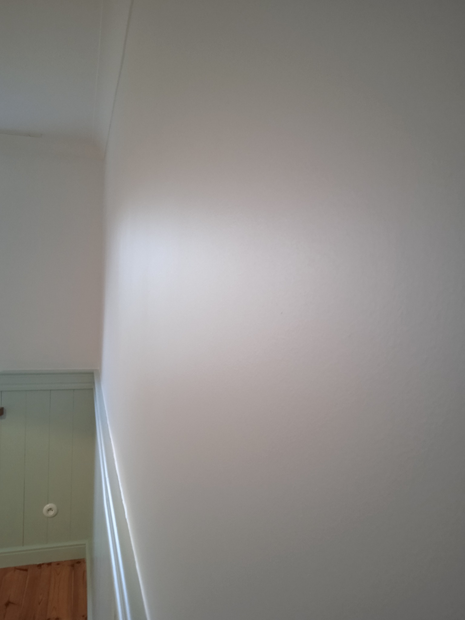 Świeżo pomalowana na biało ściana klatki schodowej, widoczna listwa przysufitowa i fragment korytarza z boazerią w kolorze pistacjowym oraz drewnianą podłogą.