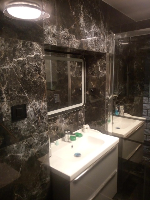 Błyszcząca, ciemna łazienka z marmurowymi płytkami, białą umywalką z szarą szafką, podświetlanym lustrem i czarnym włącznikiem światła na ścianie.