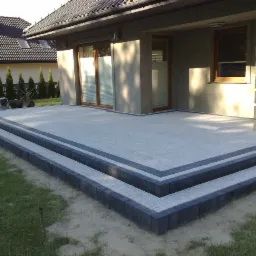 Nowoczesny taras z dwoma stopniami wykonanymi z jasnoszarej kostki brukowej obramowanej ciemnoszarą kostką, przylegający do ściany domu z oknami i kolumną z bloków betonowych.