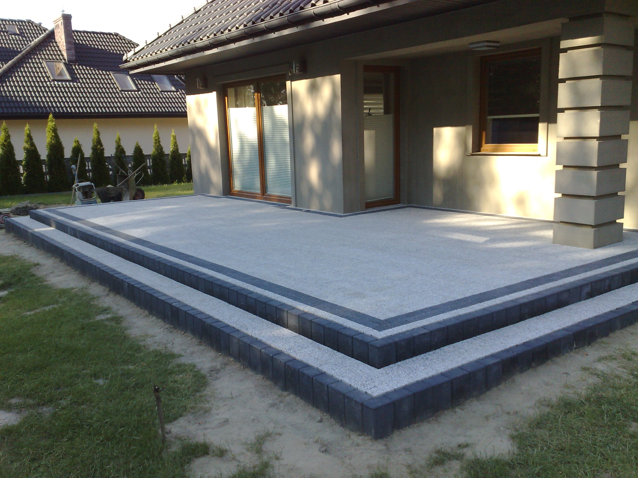 Nowoczesny taras z dwoma stopniami wykonanymi z jasnoszarej kostki brukowej obramowanej ciemnoszarą kostką, przylegający do ściany domu z oknami i kolumną z bloków betonowych.