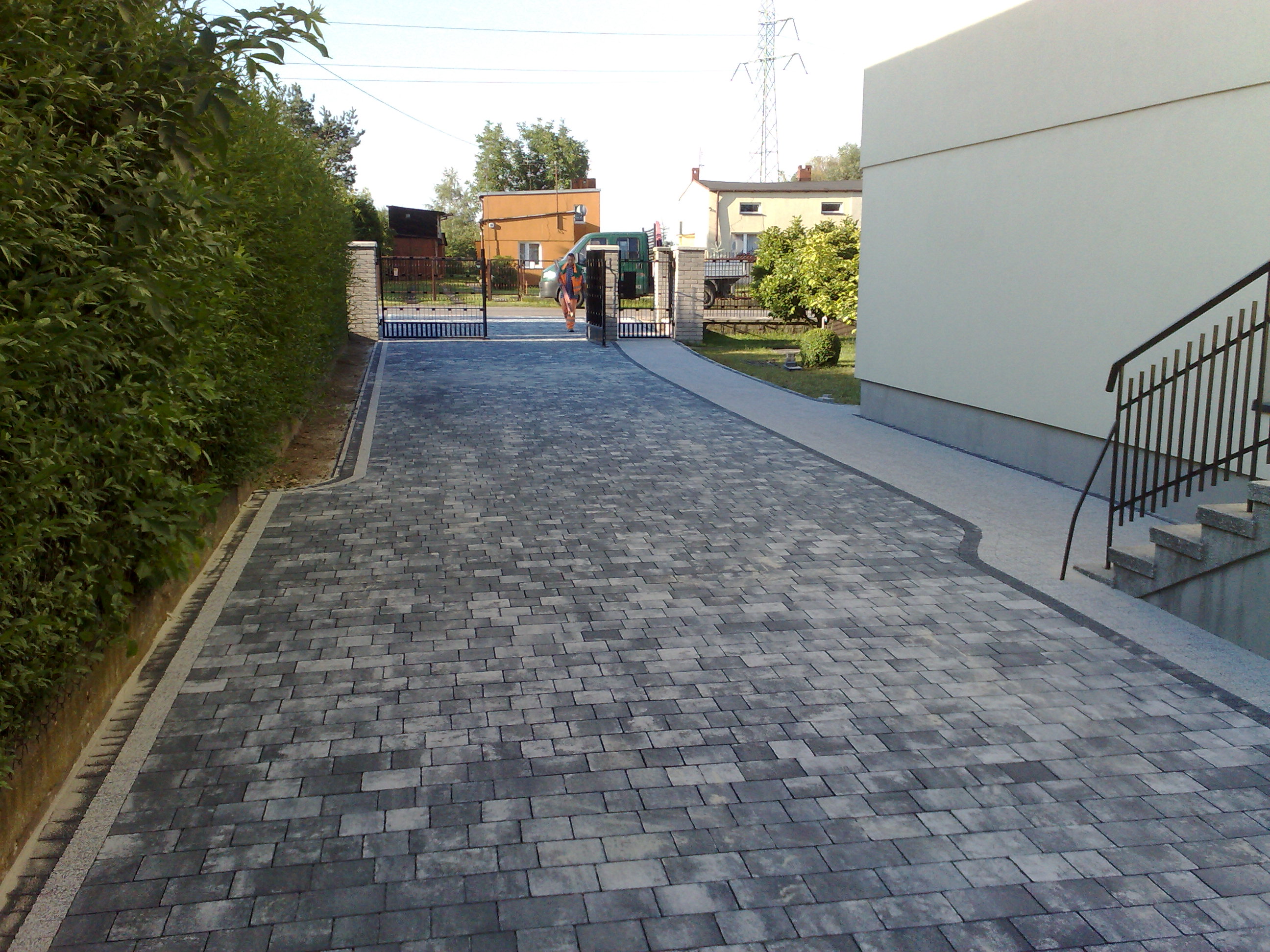 Szeroki podjazd z szarej kostki brukowej prowadzący do bramy wjazdowej, obrzeża z jasnego kamienia, po lewej stronie wysoki żywopłot, w tle budynki mieszkalne i słup wysokiego napięcia.