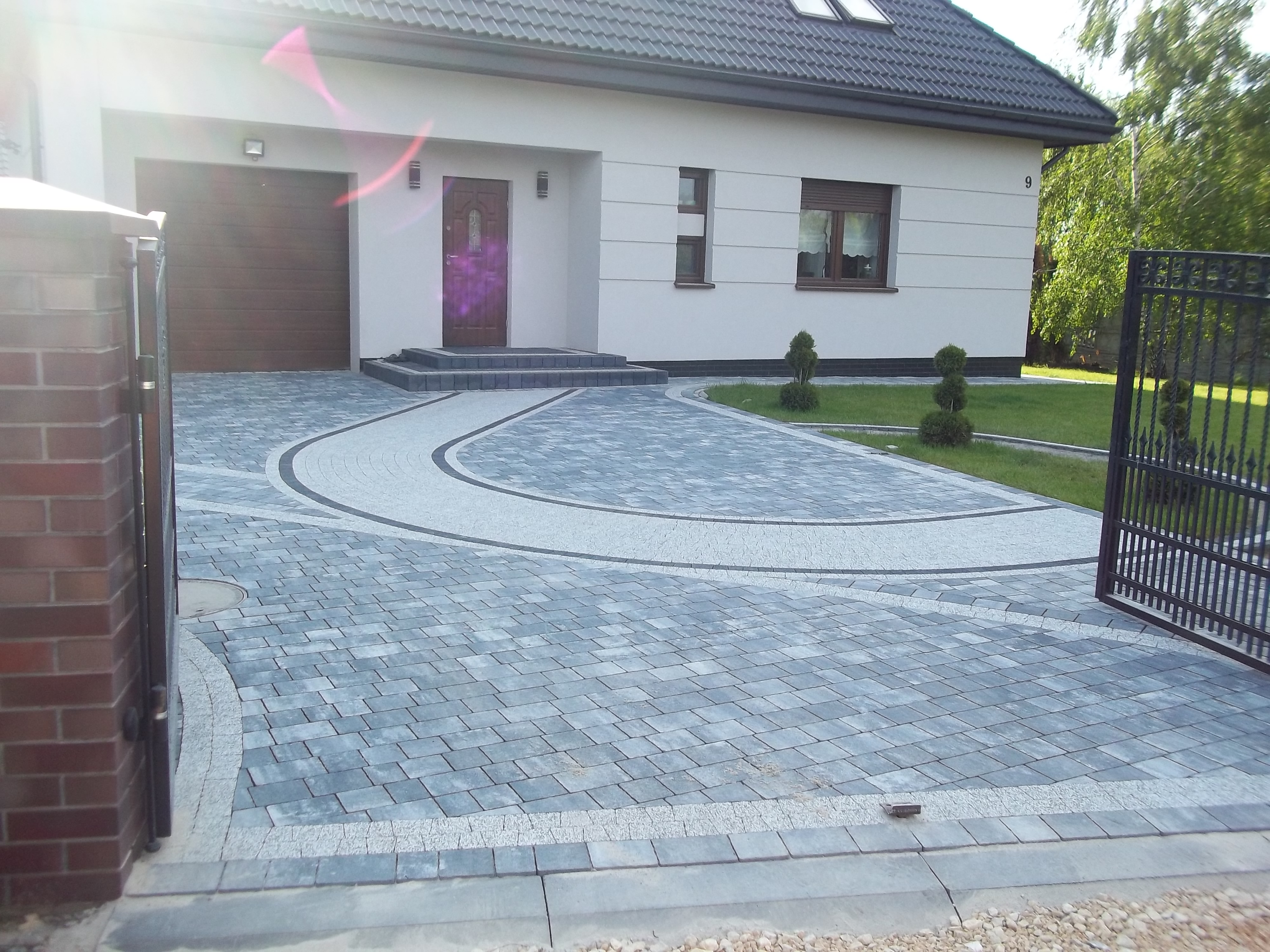 Nowoczesny podjazd z kostki brukowej w Częstochowie, z dekoracyjnym wzorem w kształcie łuku, prowadzący do domu z brązowymi drzwiami i bramą garażową, widoczny fragment metalowej bramy wjazdowej.