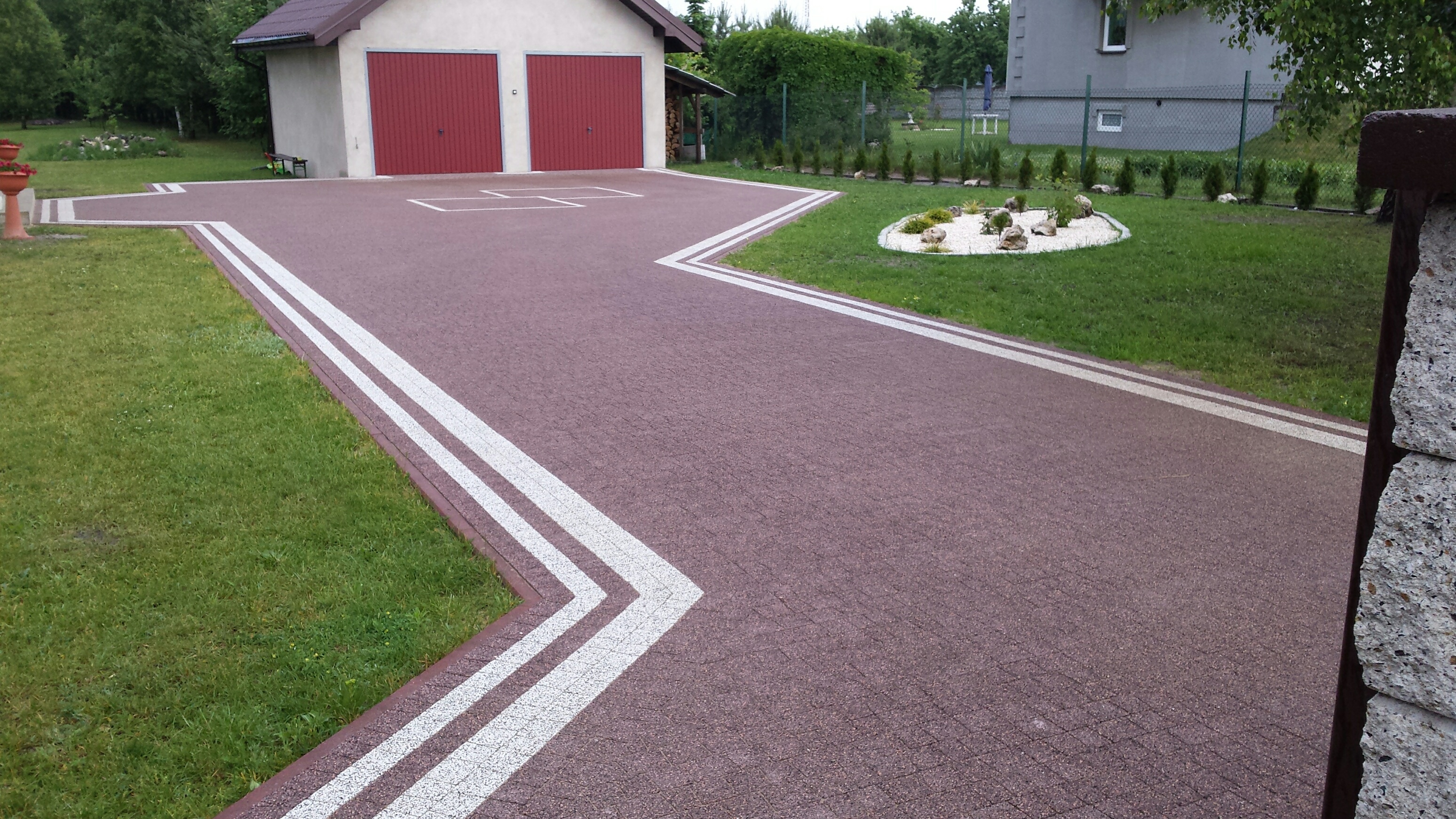 Ciemnobrązowa nawierzchnia z kostki brukowej z białymi pasami wyznaczającymi ścieżkę, prowadząca do garażu z czerwonymi drzwiami, obok trawnik z ozdobnym klombem z kamieniami i zielenią.