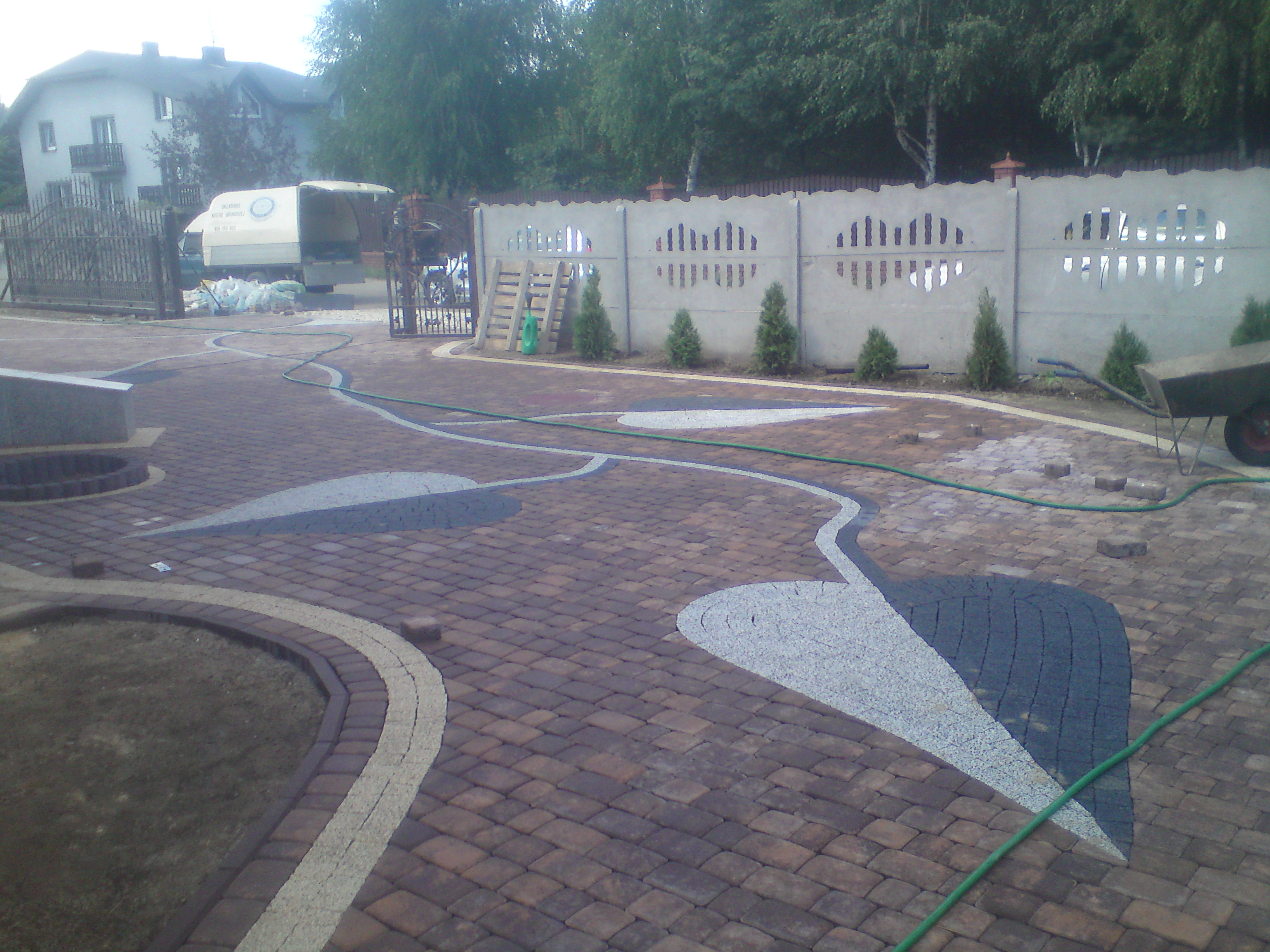 Ułożona kostka brukowa w odcieniach brązu i szarości tworząca wzory na podjeździe, z widocznym wężem ogrodowym, ogrodzeniem betonowym i młodymi drzewkami w tle.