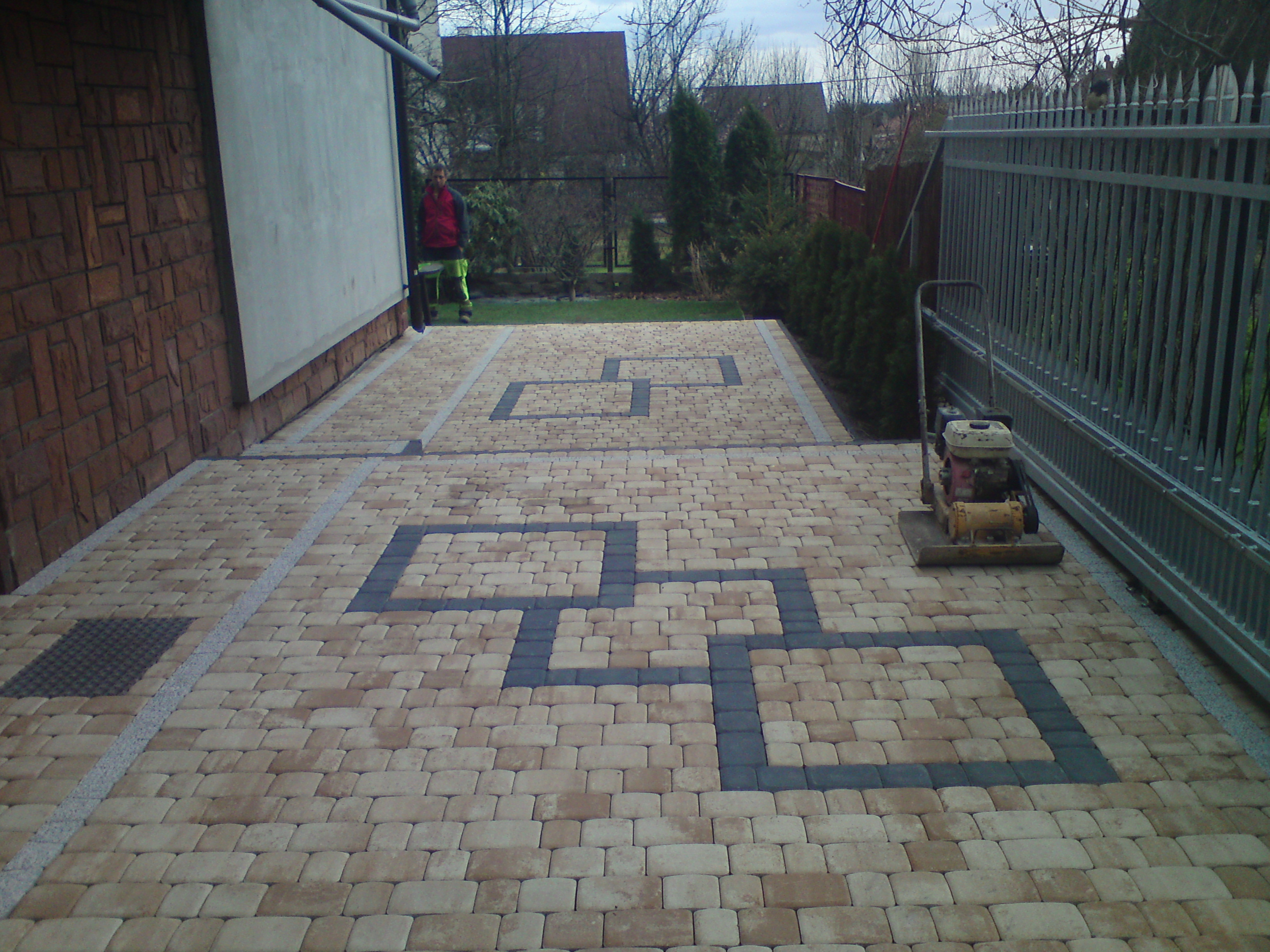 Ułożona kostka brukowa w geometryczny wzór przed domem, widoczna ubijarka wibracyjna i pracownik w tle.