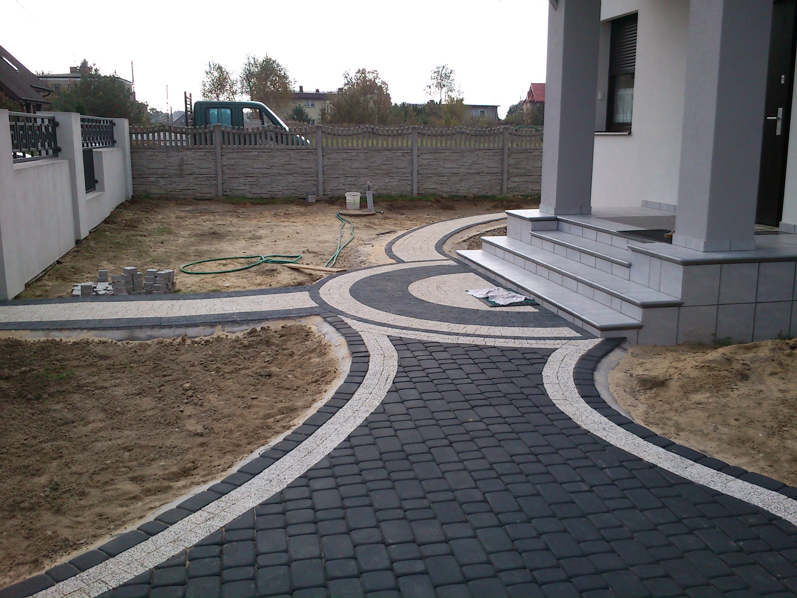 Ścieżka z ciemnej kostki brukowej z jasnymi obrzeżami prowadząca do wejścia domu, widoczne prace wykończeniowe, ziemia, węże ogrodowe i paleta kostki w tle.