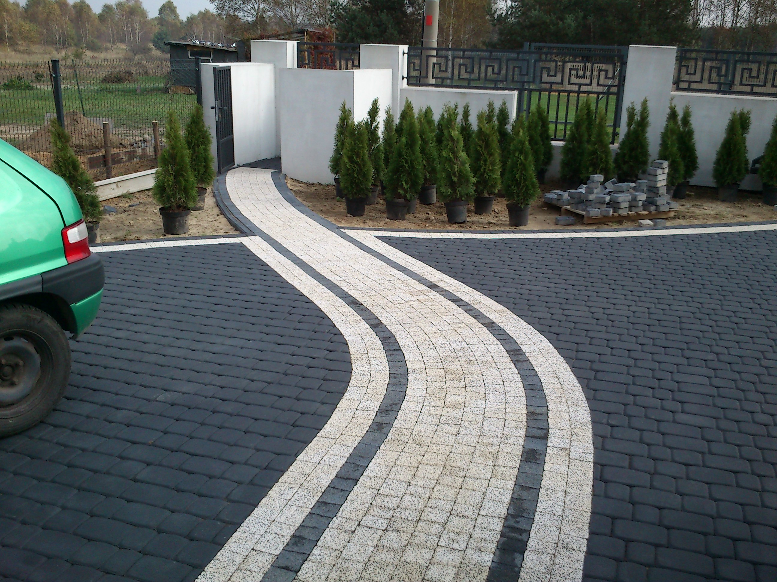 Kręta ścieżka z jasnej kostki brukowej na tle ciemniejszej nawierzchni, obok rząd tuj w donicach i zapas kostki, w tle ogrodzenie i fragment zielonego samochodu.