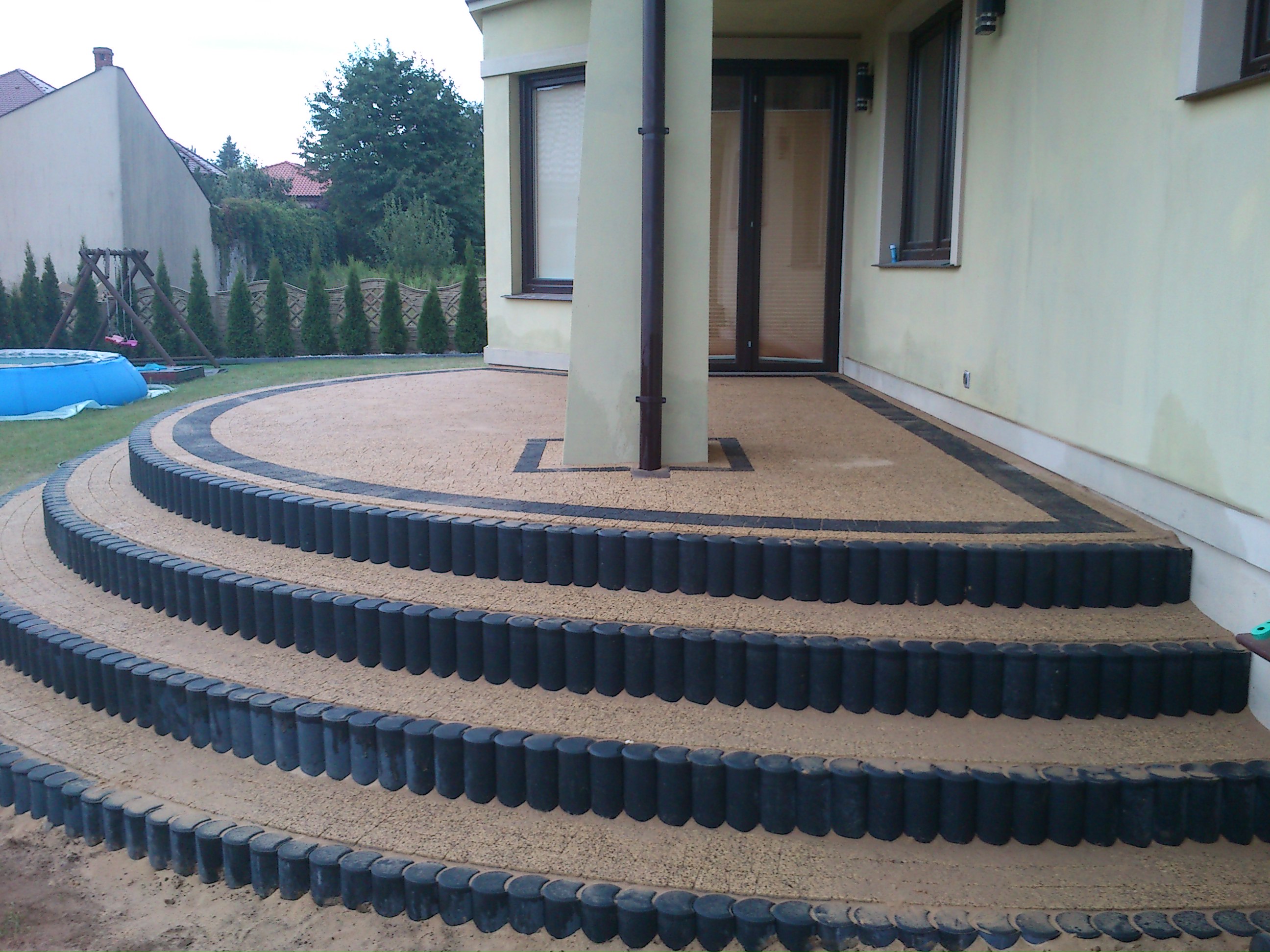 Trzystopniowe schody z kostki brukowej w kolorze piaskowym, obrzeża z czarnych elementów, prowadzące na taras przy domu z beżową elewacją; w tle trawnik, huśtawka i basen.
