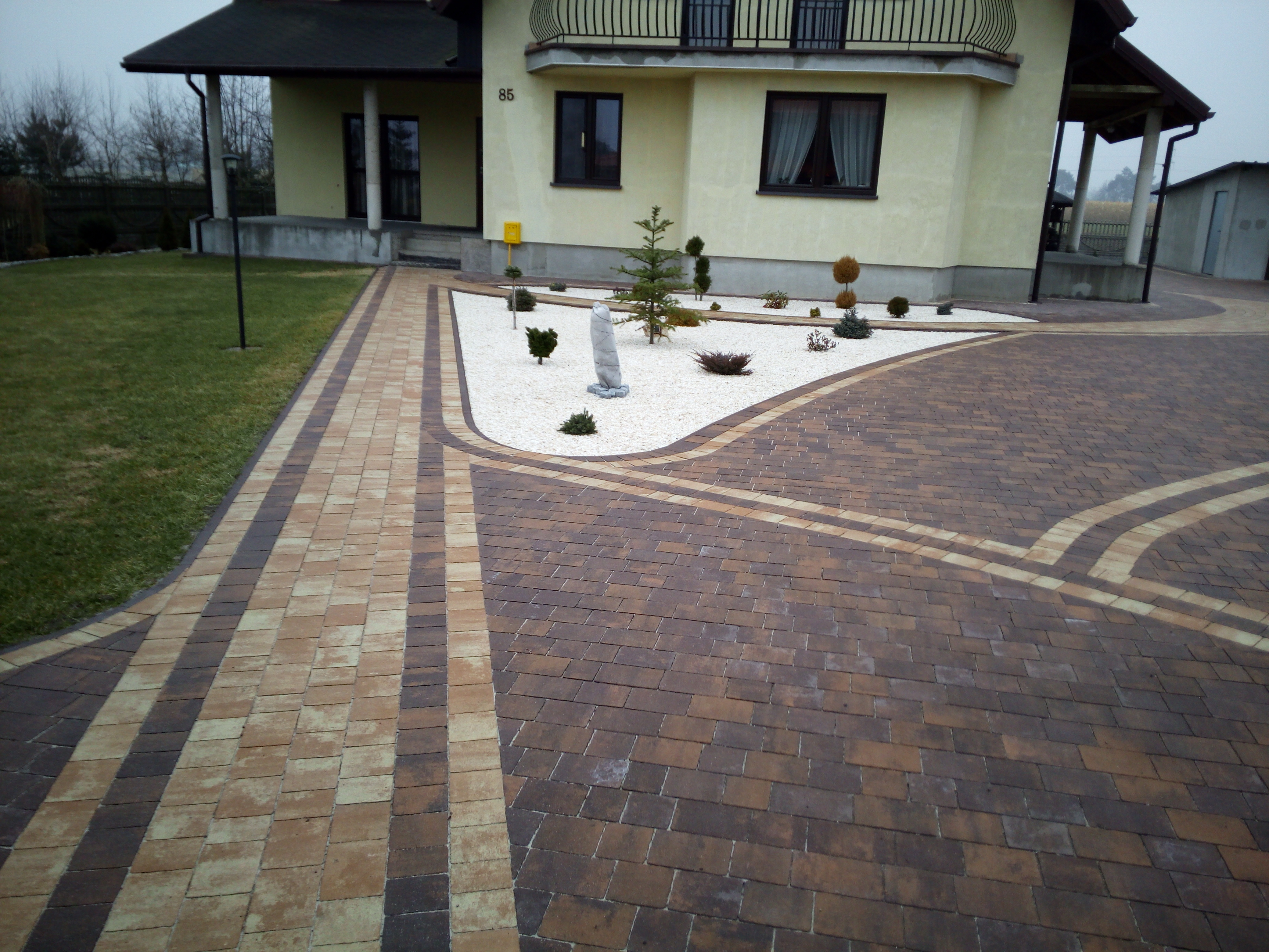 Ułożona kostka brukowa w dwóch odcieniach prowadząca do domu z numerem 85, rabata z białym kamieniem, drzewkami i rzeźbą.
