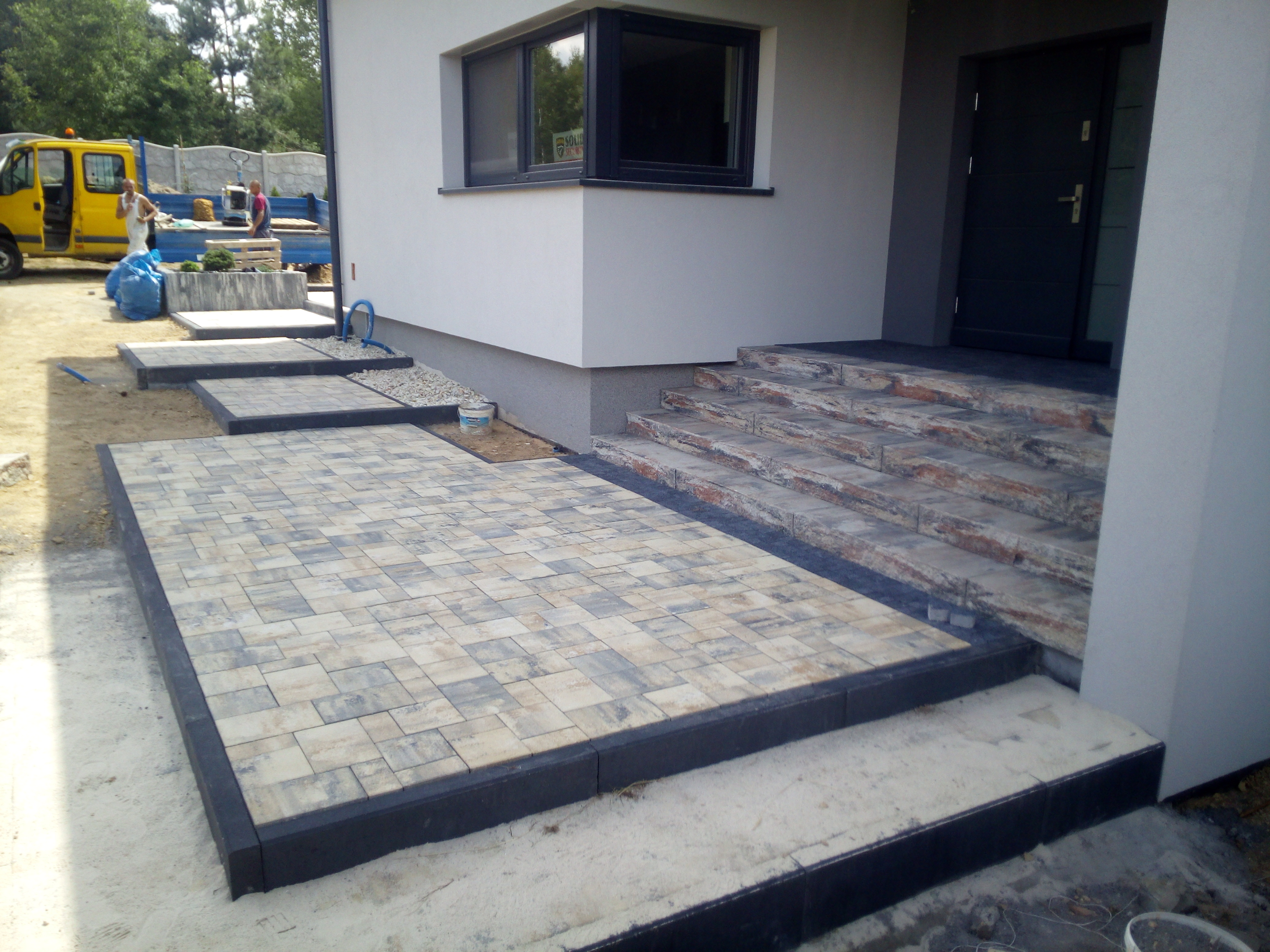 Nowoczesne wejście do domu w trakcie wykańczania: ułożona kostka brukowa w odcieniach szarości i beżu, betonowe schody z kamiennymi wstawkami prowadzące do ciemnych drzwi wejściowych, widoczny...