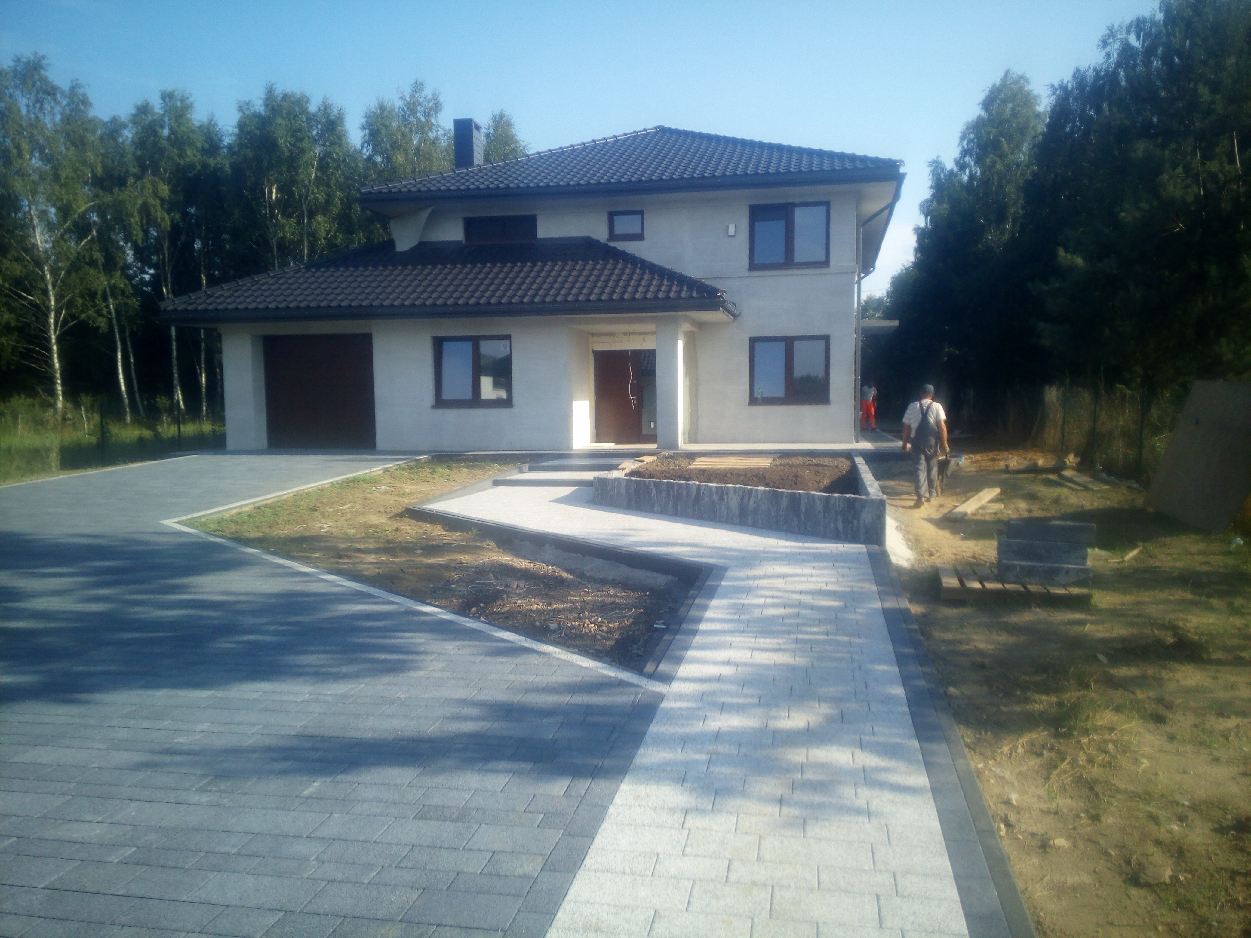 Nowoczesny dom z szarą kostką brukową na podjeździe i chodniku, pracownik w oddali, przygotowane rabaty ziemne.