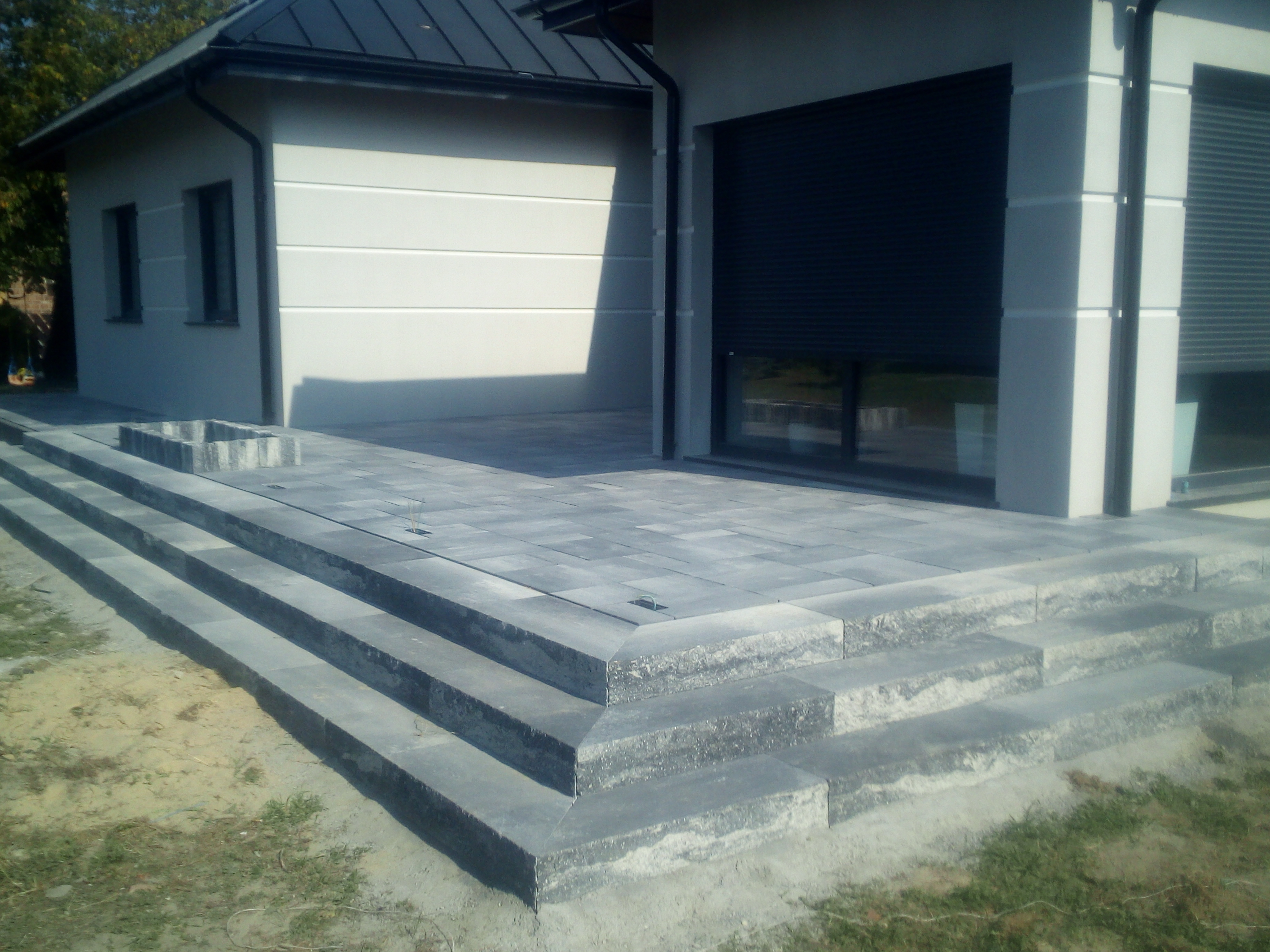 Nowoczesne schody i taras z szarej kostki brukowej prowadzące do budynku z roletami zewnętrznymi, widoczne elementy kanalizacji deszczowej w kostce.