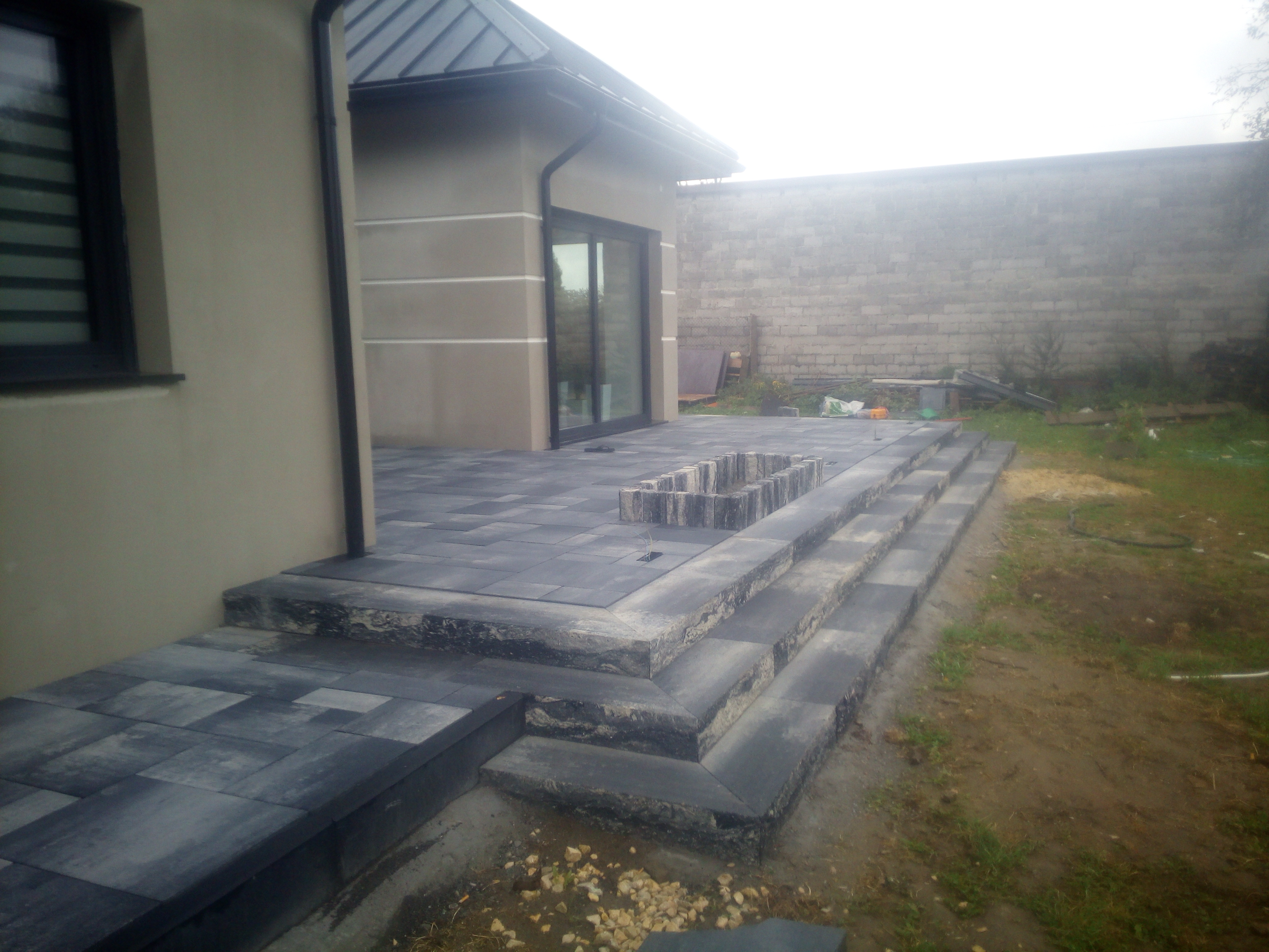 Nowoczesny taras z kostki brukowej w odcieniach szarości, z niskimi schodami i obrzeżem pod donicę, na tle jasnej elewacji domu i murowanego ogrodzenia.