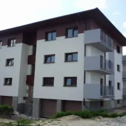 Trzy kondygnacyjny budynek z balkonami i brązowymi oknami, widziany z perspektywy trawnika. Elewacja w kolorze białym i brązowym.