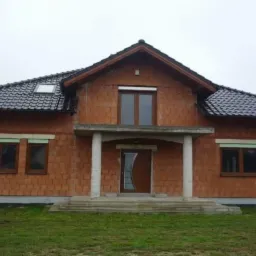 Dom w trakcie budowy z ciemną dachówką, widoczne cegły, okna z roletami i częściowo zamontowane drzwi wejściowe. Pochmurne niebo w tle.