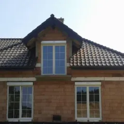 Dom w stanie surowym z trzema oknami z roletami zewnętrznymi. Widoczne palety z materiałami budowlanymi i dach pokryty dachówką.