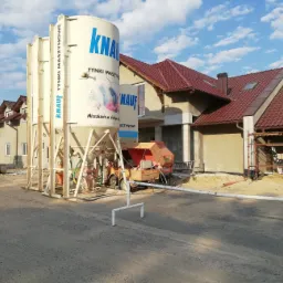 Duży silos z logo Knauf na placu budowy, obok agregat tynkarski, w tle budynek z czerwonym dachem przed tynkowaniem.