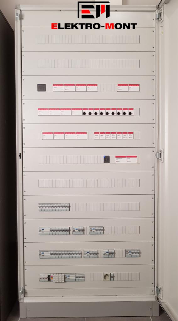 Otwarta, duża, szara szafa rozdzielcza z różnymi zabezpieczeniami, wyłącznikami i gniazdami, z logo firmy Elektro-Mont w górnej części.