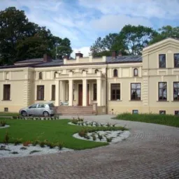 Odnowiony, kremowy dworek z kolumnami przy wejściu, otoczony zadbanym trawnikiem i brukowanym podjazdem, z zaparkowanym samochodem osobowym.