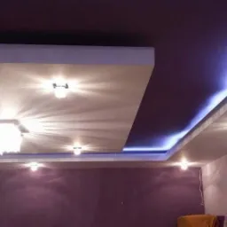 Dekoracyjny sufit podwieszany z oświetleniem LED i centralnym żyrandolem, fioletowe ściany w tle.