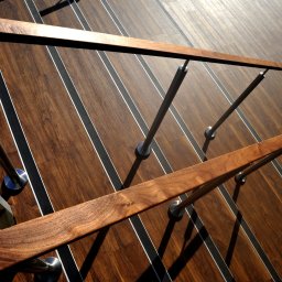 Opiela-parkiety.pl - Nowoczesne schody z ciemnego drewna z metalowymi poręczami i minimalistycznymi detalami, oświetlone mocnym światłem słonecznym, tworzącym wyraźne cienie.