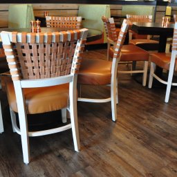 Opiela-parkiety.pl - Krzesła z plecionymi oparciami i brązowymi siedziskami na drewnianej podłodze. Widoczne stoły i zielone kanapy w tle. Ujęcie z niskiego kąta.