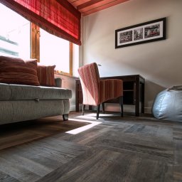 Opiela-parkiety.pl - Ułożona w jodełkę podłoga z paneli w salonie z szarą sofą, krzesłem w paski i workiem sako. Promienie słońca padają na podłogę.