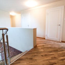 Opiela-parkiety.pl - Jasne, drewniane panele podłogowe ułożone w korytarzu z białymi drzwiami i schodami z metalową balustradą w Otwocku. Widok na ceglaną ścianę na schodach.