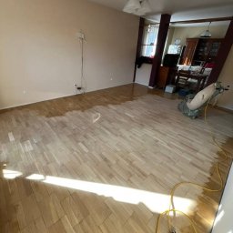 Opiela-parkiety.pl - Cyklinowanie parkietu w trakcie realizacji. Część podłogi już wycyklinowana, widoczna maszyna do cyklinowania i kabel zasilający. Jasne drewno, naturalne światło.