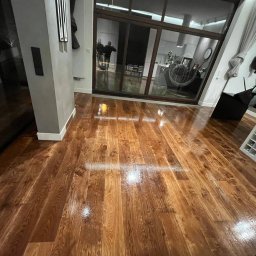 Opiela-parkiety.pl - Błyszcząca, świeżo polakierowana podłoga z desek w odcieniach brązu, odbijająca światło. Widoczne duże okno przesuwne i fragmenty wyposażenia wnętrza.