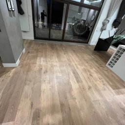 Opiela-parkiety.pl - Jasna, drewniana podłoga ułożona w salonie z dużym oknem przesuwnym. Widoczny fragment kolumny i mebli. Naturalne oświetlenie podkreśla strukturę drewna.