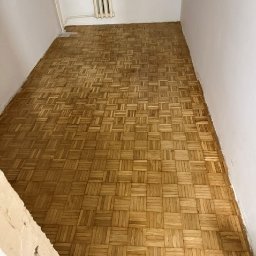 Opiela-parkiety.pl - Wnętrze z odnowioną, jasną podłogą parkietową w geometryczny wzór. Widoczne ślady po cyklinowaniu, grzejnik i fragment ściany w trakcie remontu.