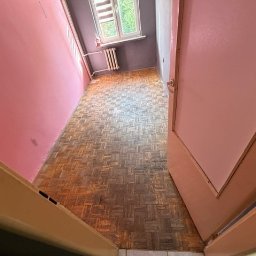 Opiela-parkiety.pl - Pokój z parkietem w jodełkę przed cyklinowaniem. Widoczne zniszczenia i przebarwienia na podłodze. Różowe ściany, okno z grzejnikiem w tle. Ujęcie zza otwartych drzwi.