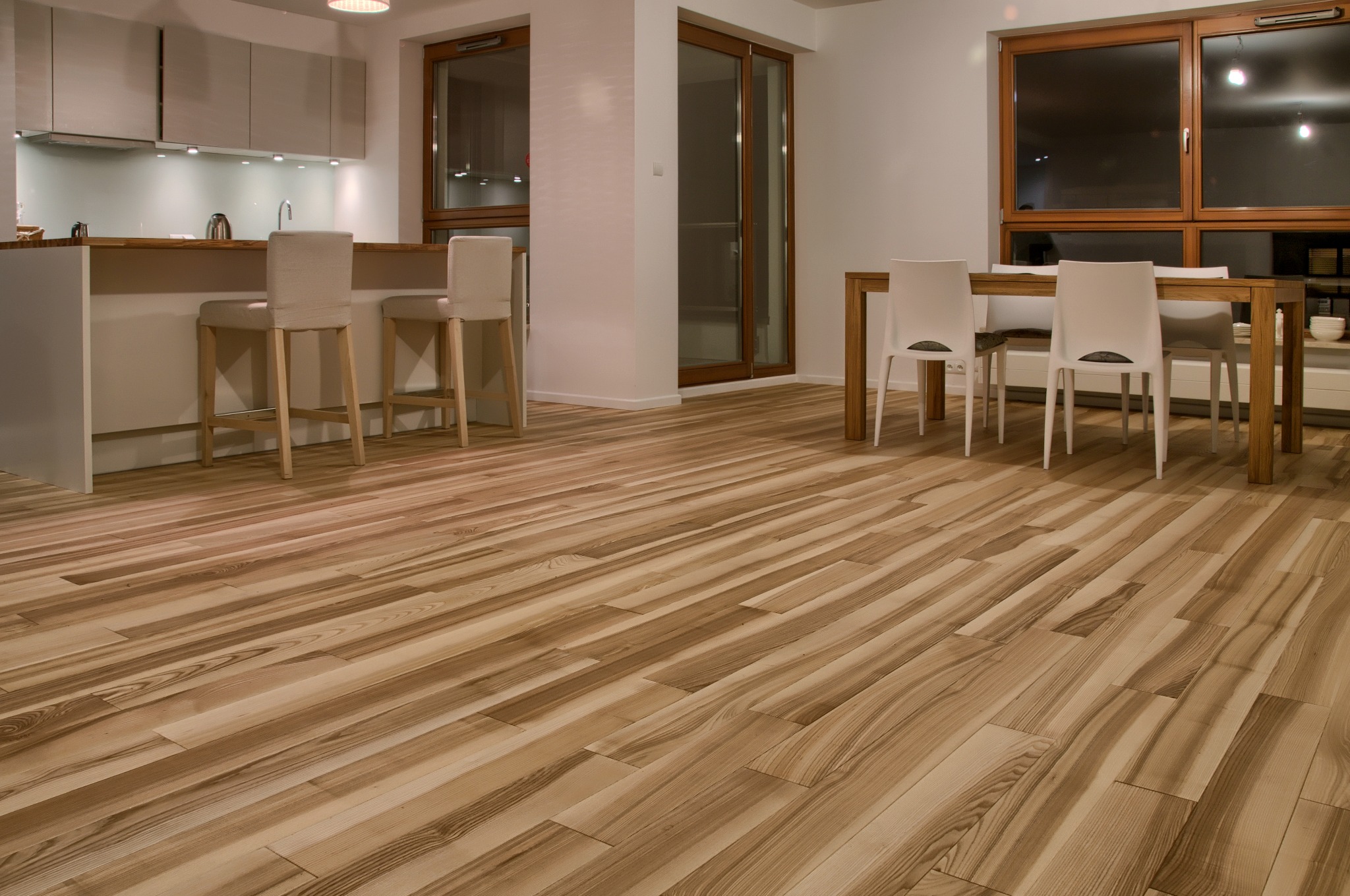 Jasna, drewniana podłoga ułożona w jadalni z aneksem kuchennym. Widoczne krzesła barowe i stół z krzesłami. Ciepłe oświetlenie wnętrza.