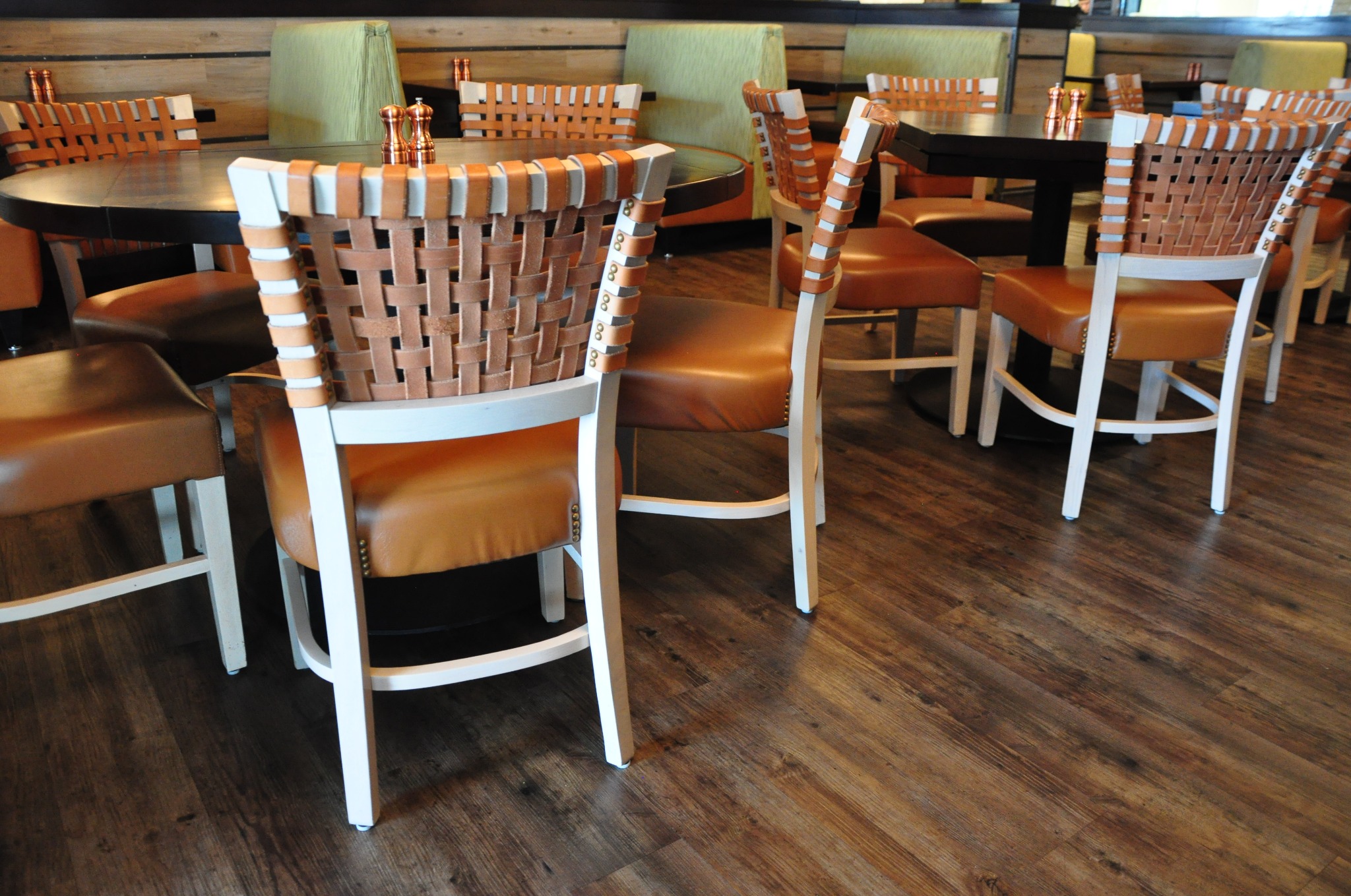 Krzesła z plecionymi oparciami i brązowymi siedziskami na drewnianej podłodze. Widoczne stoły i zielone kanapy w tle. Ujęcie z niskiego kąta.