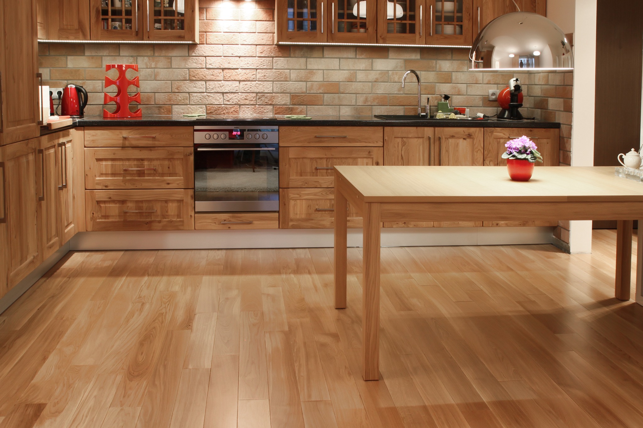 Jasna, drewniana podłoga parkietowa w kuchni z drewnianymi szafkami i ceglaną ścianą. Na stole kwiaty w doniczce. Widoczne oświetlenie podszafkowe.