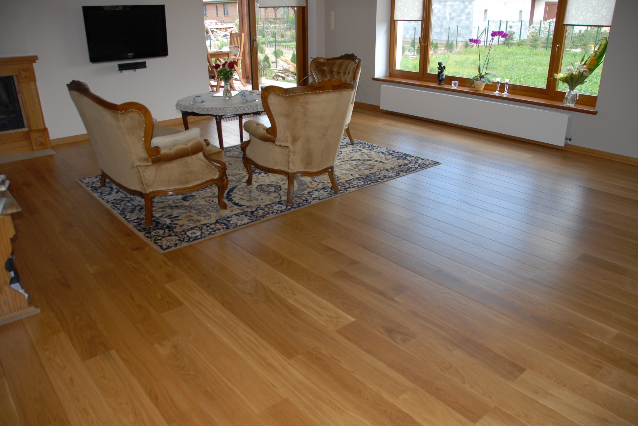 Jasna, drewniana podłoga z parkietu ułożonego w jodełkę, z widocznymi meblami w tle, dywanem i oknem z widokiem na ogród. Ciepłe oświetlenie wnętrza.