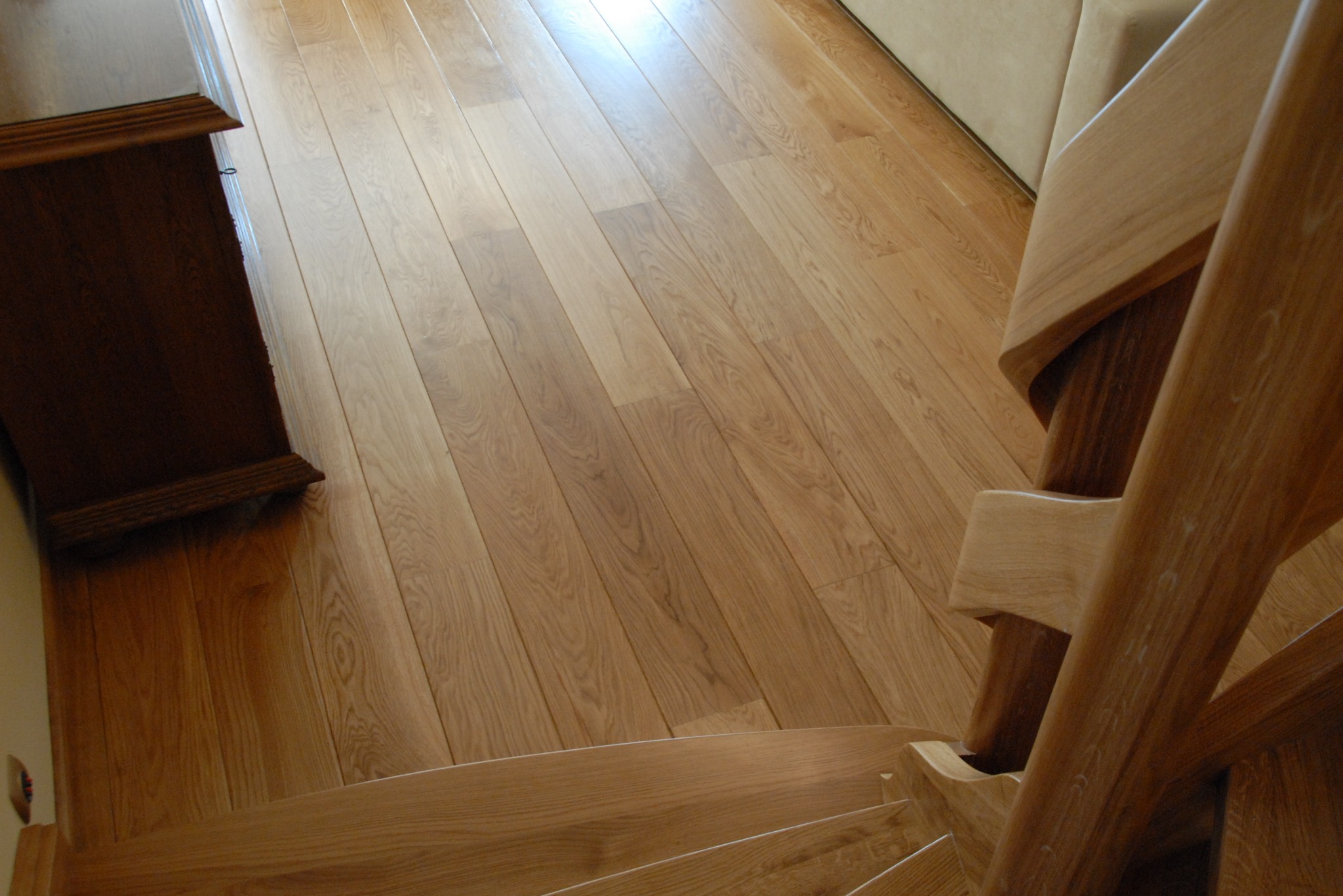 Jasny parkiet ułożony w jodełkę francuską, widok z góry na drewniane schody z balustradą. Fragment szafki przy ścianie. Naturalne światło.