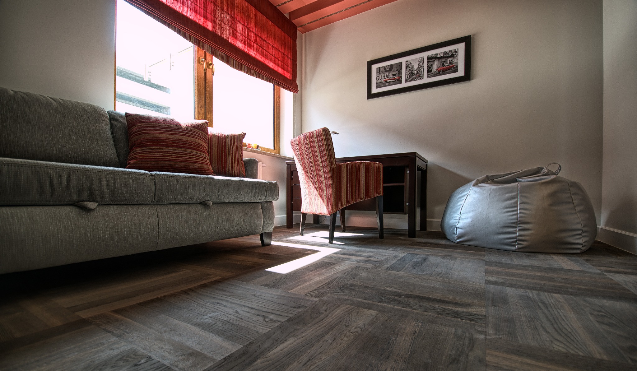 Ułożona w jodełkę podłoga z paneli w salonie z szarą sofą, krzesłem w paski i workiem sako. Promienie słońca padają na podłogę.