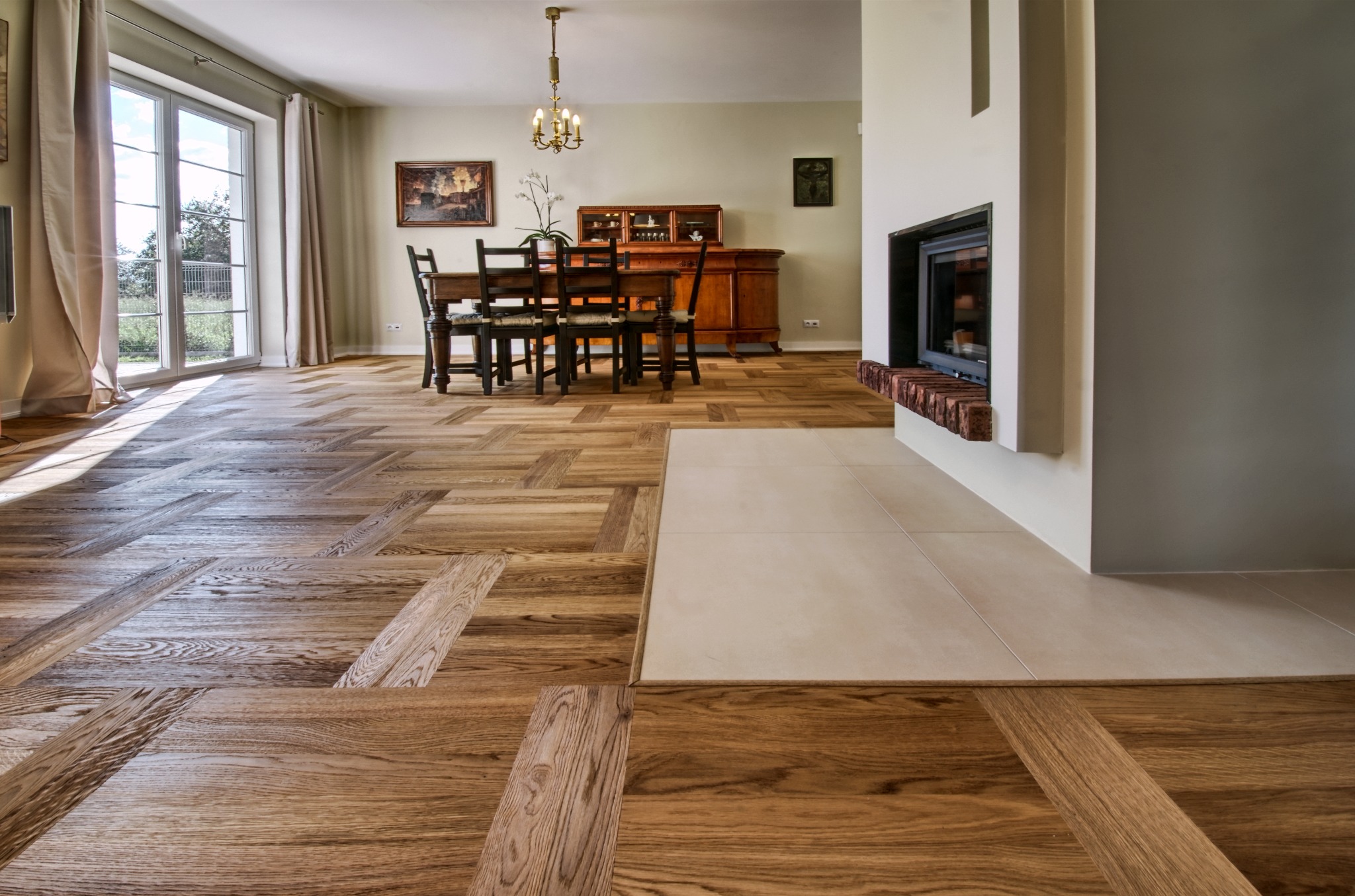 Parkiet ułożony we wzór jodełki francuskiej w salonie z kominkiem. Jasne drewno, widoczne słoje. W tle stół z krzesłami i kredens. Duże okno z widokiem na zewnątrz.