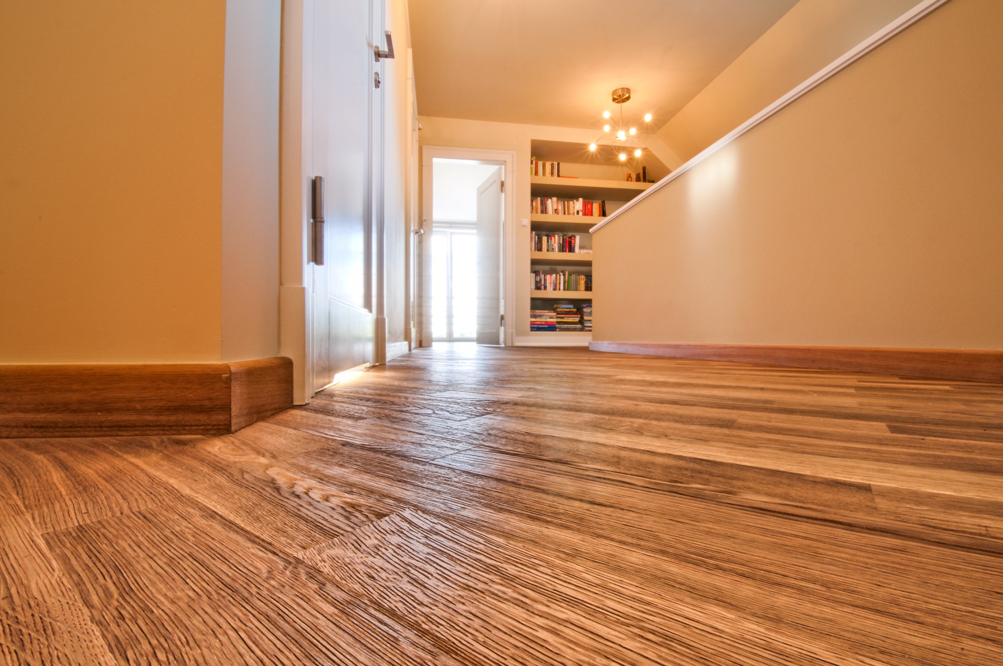 Jasne panele podłogowe z wyraźną fakturą drewna, widok z niskiej perspektywy, fragment ściany i drzwi w tle, ciepłe oświetlenie wnętrza.