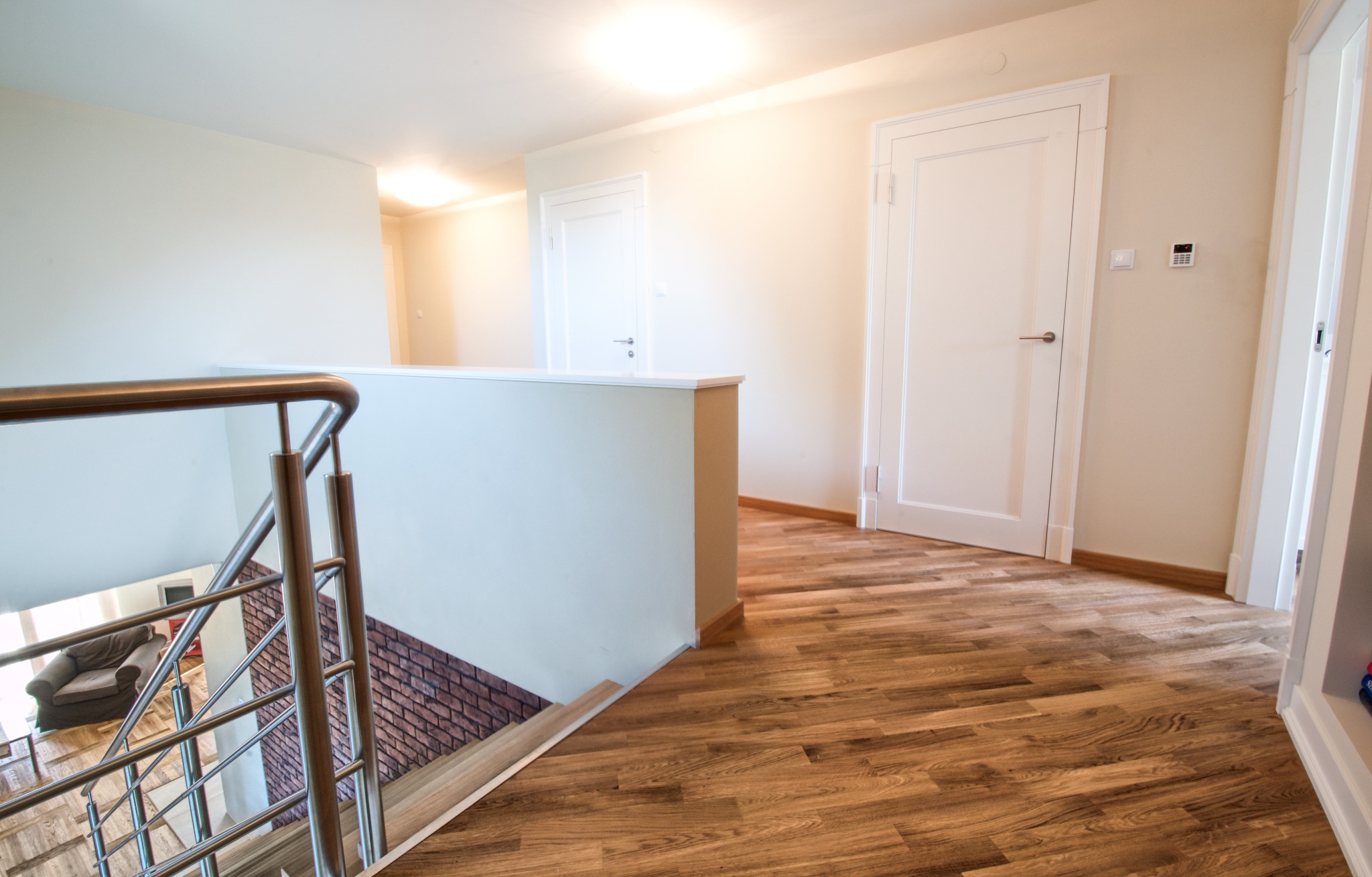 Jasne, drewniane panele podłogowe ułożone w korytarzu z białymi drzwiami i schodami z metalową balustradą w Otwocku. Widok na ceglaną ścianę na schodach.