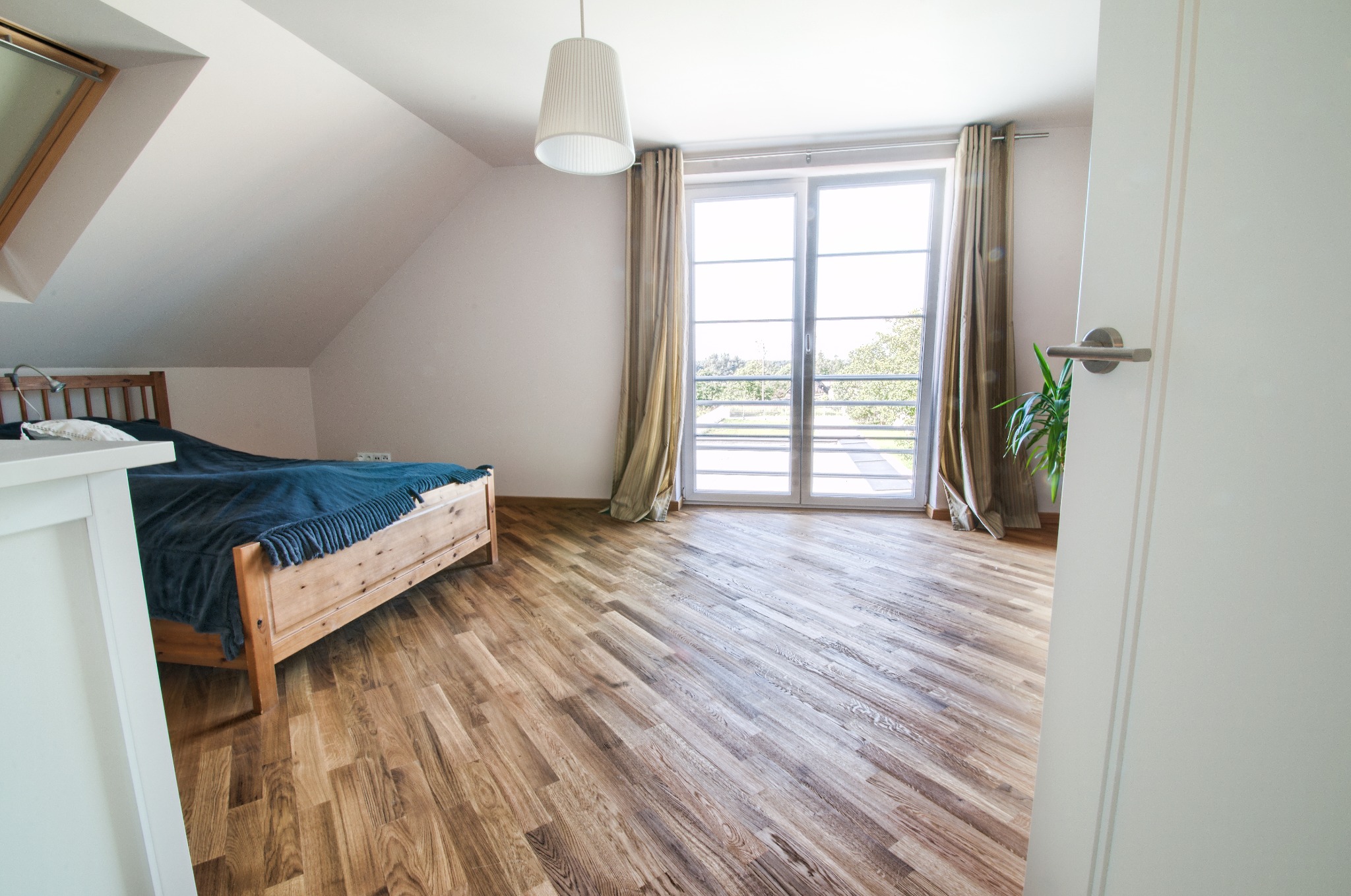 Jasne poddasze z drewnianą podłogą ułożoną w jodełkę, łóżko z niebieską narzutą, okno balkonowe z zasłonami i białe drzwi z klamką.