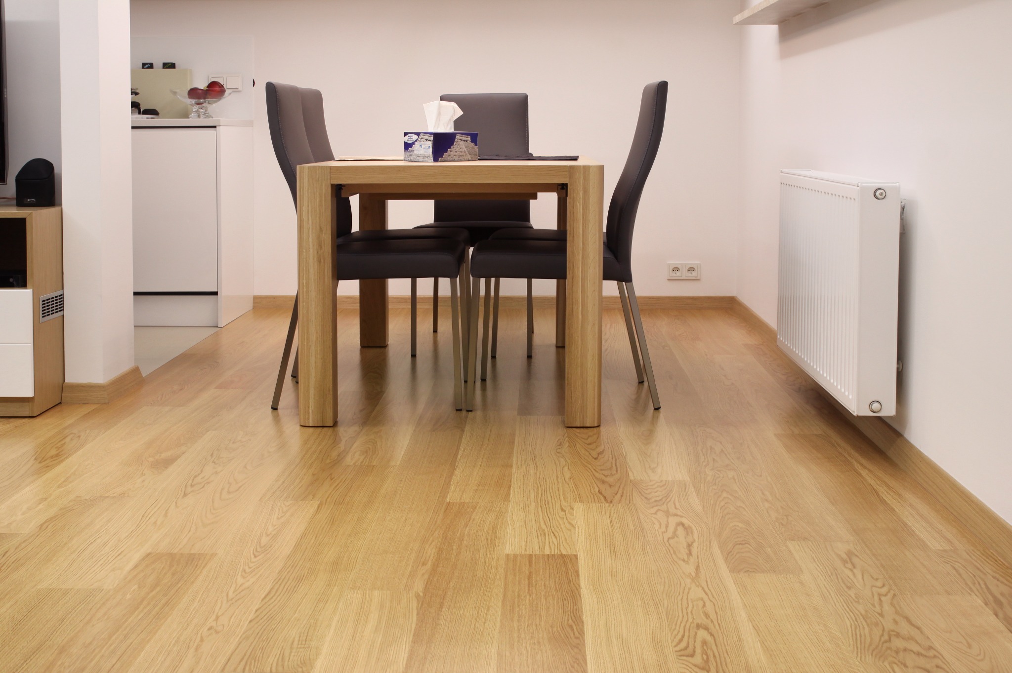 Jasne panele podłogowe w jadalni z drewnianym stołem i szarymi krzesłami. Widoczny biały grzejnik i fragment kuchni w tle. Ujęcie z niskiej perspektywy.