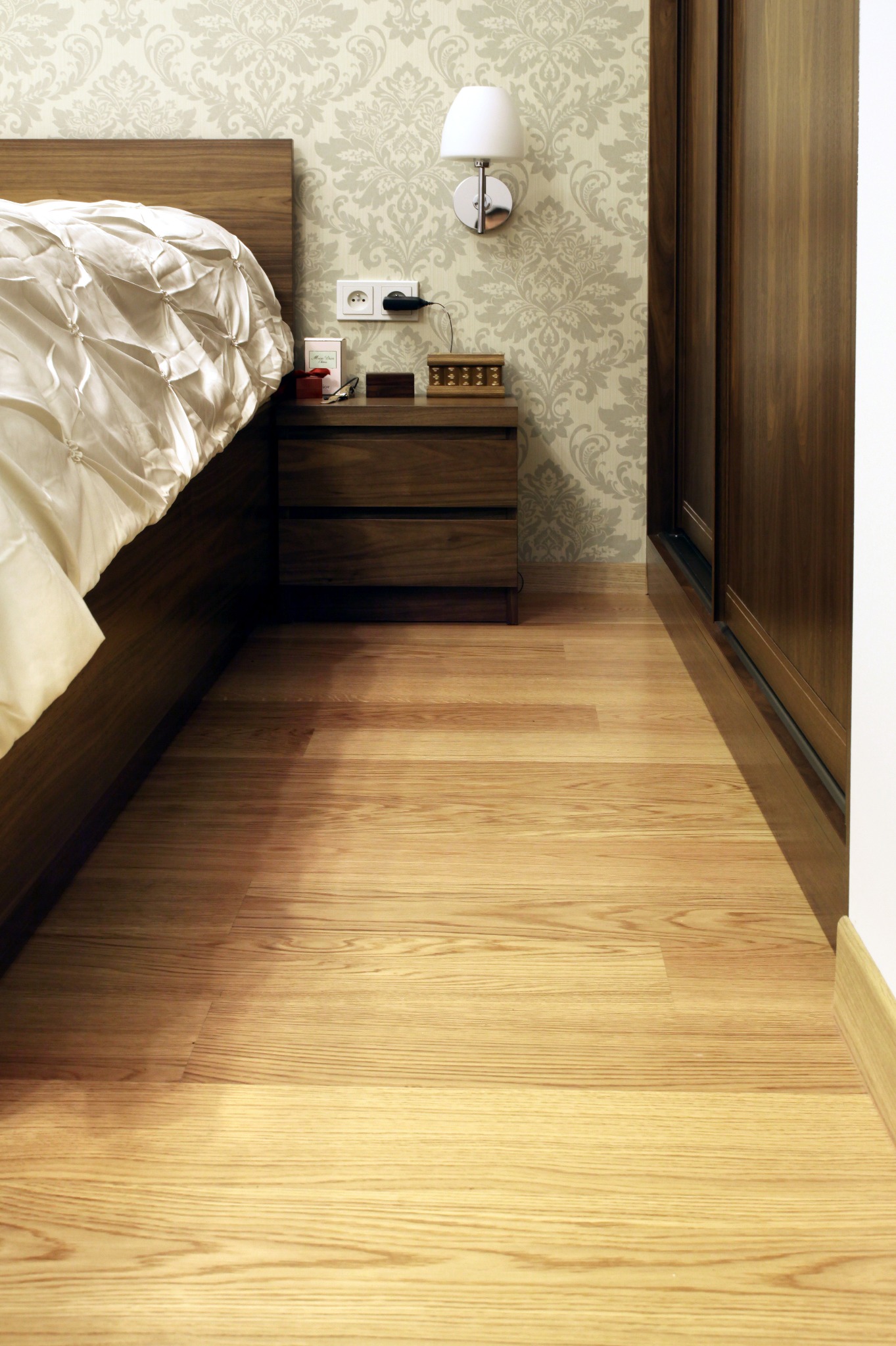 Jasna podłoga z paneli drewnopodobnych w sypialni, widok z niskiej perspektywy. Fragment łóżka i szafki nocnej po lewej, szafa przesuwna po prawej.