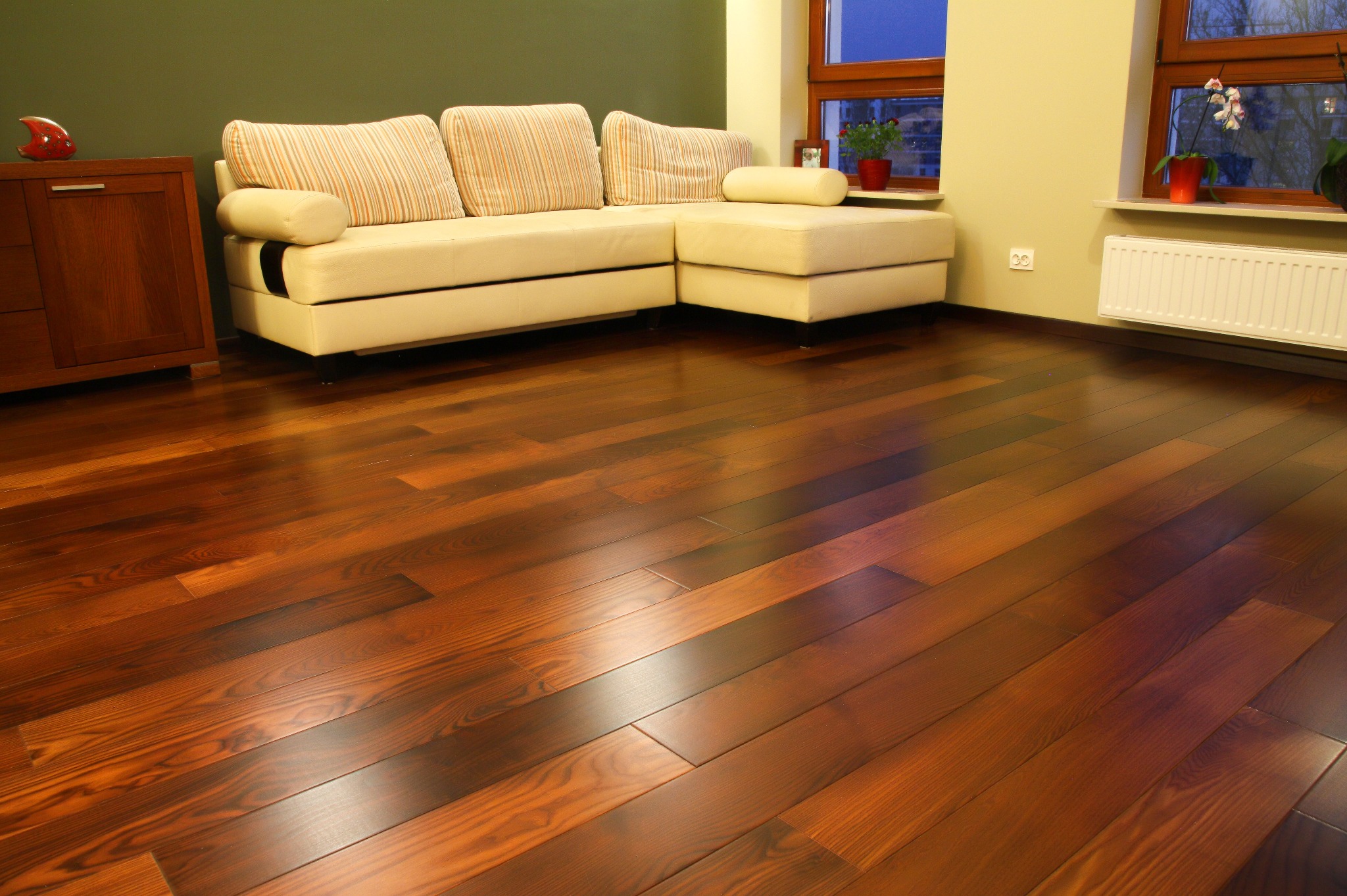 Polakierowany parkiet z egzotycznego drewna w salonie z beżową sofą i zieloną ścianą. Widoczne okno z kwiatami i grzejnik.