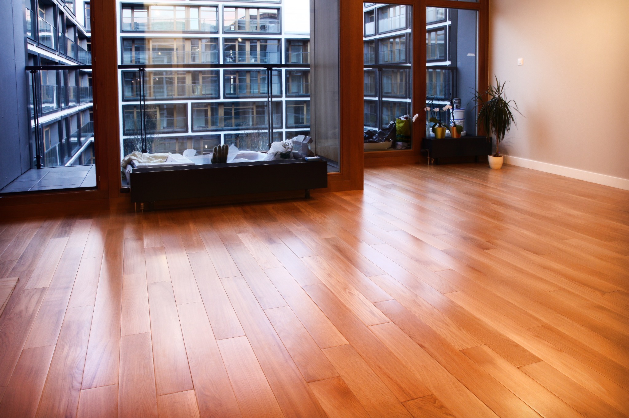 Jasny parkiet w nowoczesnym wnętrzu z dużymi oknami balkonowymi. Widok na budynek z balkonami. Minimalistyczny wystrój z roślinami doniczkowymi.