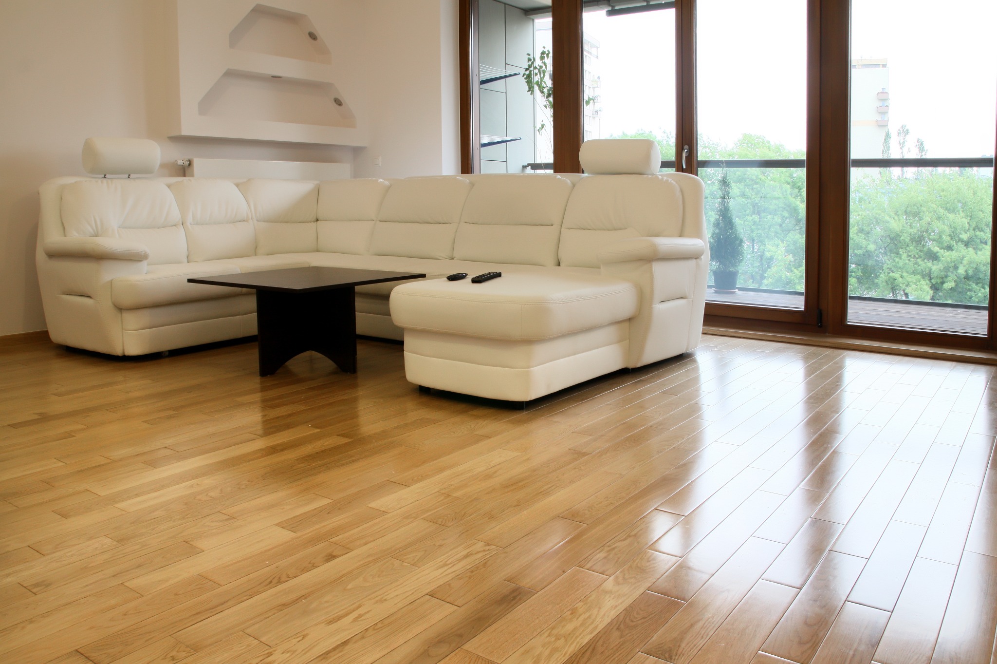 Jasny parkiet w salonie z białym narożnikiem i dużym oknem balkonowym. Parkiet ułożony w jodełkę, widoczne refleksy światła. Dwa piloty leżą na narożniku.